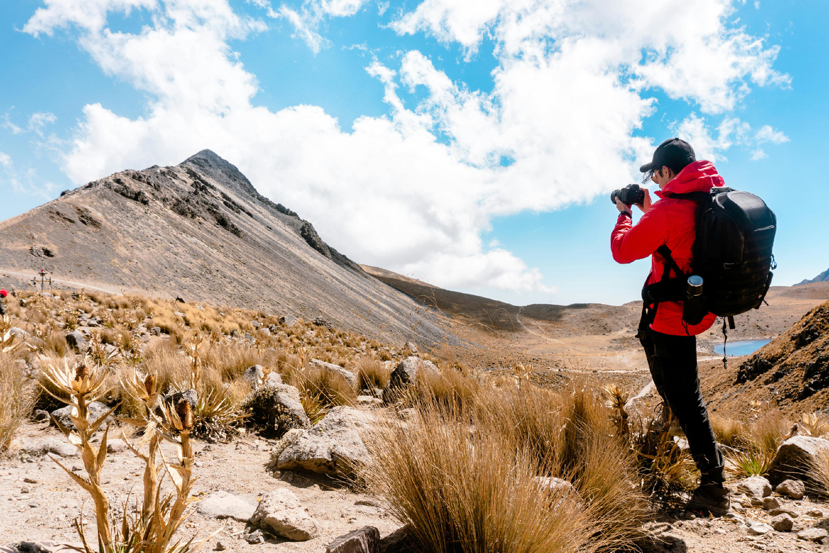Путешественник в красной куртке с рюкзаком фотографирует горный пейзаж на фоне голубого неба с облаками