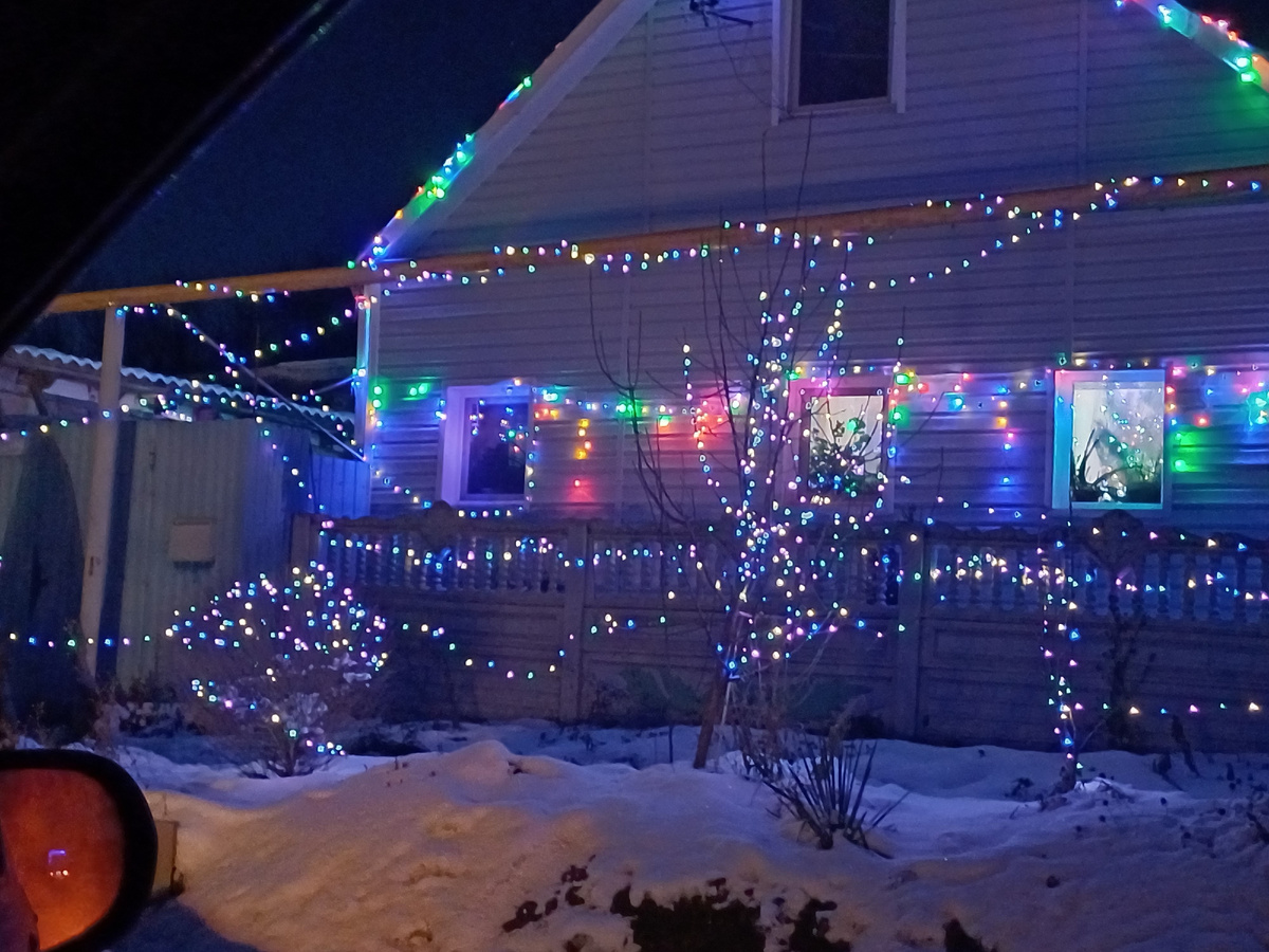 Гирляндами обвит дом и все деревья. На фотографии не заметно, а в жизни гирлядны развешаны по чётким линиям. Спереди мелкие часто мигающие огоньки, а на заднем плане огоньки крупнее и они немного застывшие. Фото автора Юлии Солоха. 