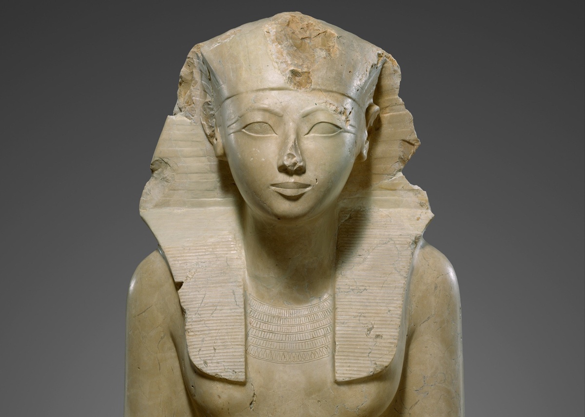 Известняковая сидячая статуя Хатшепсут в Метрополитен-музее, Нью-Йорк. Царица изображена без накладной бороды, но с символами власти фараона: головной убор немес, увенчанный царским уреем. 