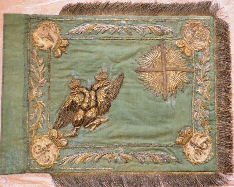 Штандарт эскадронный Астраханского кирасирского полка образца 1811 г. на древке с навершием. На штофном полотнище зеленого цвета изображен двуглавый орел. В правом верхнем углу - крест, окруженный сиянием, по углам императорский вензель Александра I, окруженный венком. Двусторонний. По верхнему, нижнему и правому краю - бахрома. Древко черного с желобками выкрашенные светлой краской, в нижней части сохранился подток (металлический «стакан» конусовидной формы, защищавший дерево), чуть выше - кожаный ремень-петля. В верхней части помещено кольцо с надписью: «1811 года Астраханский кирасирский полк 1852 год Кирасирскаго Его Императорскаго Высочества Виликаго Князя Николая Николаевича Астраханского кирасирского полка 1-го дивизиона». Навершие образца 1803 года, в форме копья, в середине помещен объемный двуглавый орел с поднятыми крыльями, под короной, в лапах - скипетр и держава, без гербового щита на груди. Ссылка: https://goskatalog.ru/portal/#/collections?id=7844004