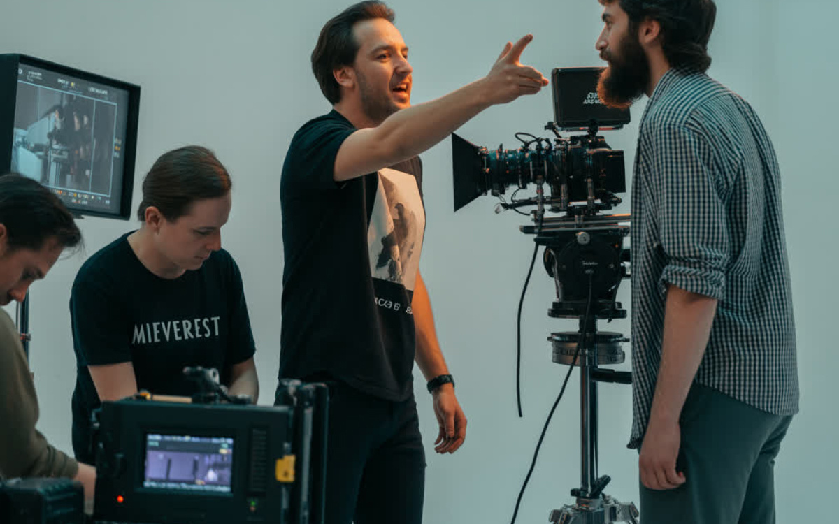 Режиссёр на съёмочной площадке даёт указания актёру, стоящему перед камерой, в то время как оператор и ассистенты контролируют процесс с мониторов.