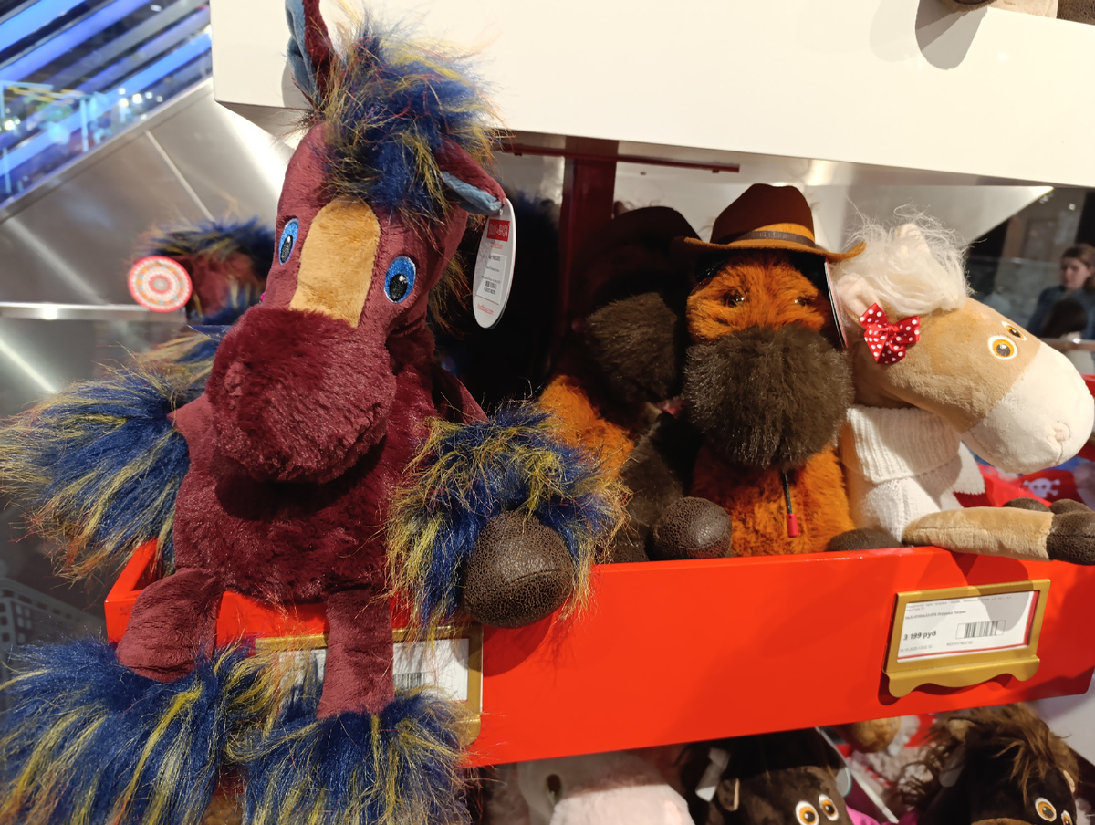 Цветная фотография горизонтальной ориентации. Стойка-этажерка с игрушками в «Центральном Детском мире». Перед нами полка оранжевая, выше – белая. На оранжевой сидят меховые лошади. Справа задумчивая светло-бежевая с белой мордочкой, белой пушистой чёлкой и в белом вязаном свитере с горлышком, лупоглазая, глазки вышиты, круглые, рыжие с коричневыми зрачками, у чёлки красный бантик в белый горошек. Рядом – явно мужчины – рыжие кони в ярко-оранжевых ковбойских шляпах. Если бы это были люди, то я дала бы им на вид лет шестьдесят – не старые, но уже умудрённые жизненным опытом, чуть уставшие, но озорные. На кончиках ног – кожаные вставки, мордочки меховые, тёмно-коричневые, глаза карие, яркие, со зрачками, пластиковые, блестят как настоящие. На углу полки, ближе к нам, сидит лошадь меховая, огненно-коричневая, чуть задумчивая, выше носа – продолговатая бежевая вставка, глаза вышитые ярко-синие с чёрными зрачками. На кончиках ног и на голове синее опушение с проблесками жёлтого мелирования, на левом ухе круглая бумажная бирка. За полкой с игрушками стеклянная перегородка, вдалеке справа две женщины.