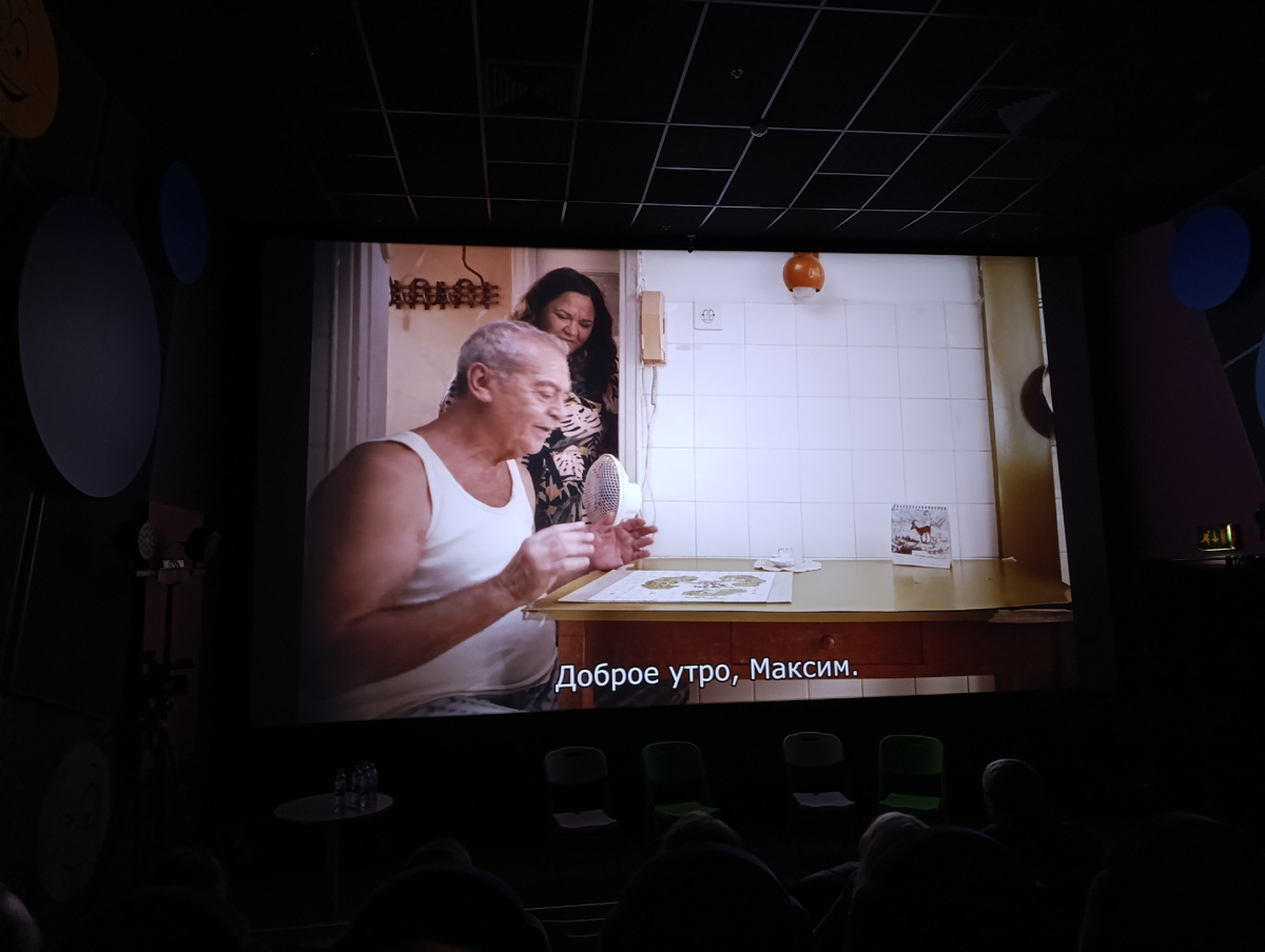 Цветная фотография горизонтальной ориентации. Небольшой тёмный кинозал. Вид слева, под углом. На первых рядах сидят зрители. На стенах круглые украшения. Около экрана в ряд поставлены четыре стула, слева на круглом столике стоят бутылки с водой. На экране цветной кадр из фильма «Анна – Максим». Обычная кухня, стена выложена белой плиткой, на ней висит телефонная трубка, рядом розетка, коричневый светильник. Около стены небольшой прямоугольный столик. На нём стоит белый вентилятор, что-то типа подсвечника и календарь на пружинке, с изображением горного козла. За столом слева на фото, боком к нам, сидит полноватый пожилой мужчина, мускулистый, загорелый, в белой майке и шортах. У него короткая стрижка, седоватые волосы, крупное лицо, широкие тёмные брови. Он слегка развёл руки и чуть удивлённо рассматривает картину, лежащую перед ним на столе (или это какой-то медицинский рисунок). Поодаль, по его левую руку, стоит молодая женщина, чуть полноватая, в чёрном халатике с листьями монстеры. Волосы тёмно-каштановые, слегка волнистые, по плечи. Тоже смотрит на изображение, лежащее на столе. Внизу экрана строка субтитров, белый шрифт: «Доброе утро, Максим».