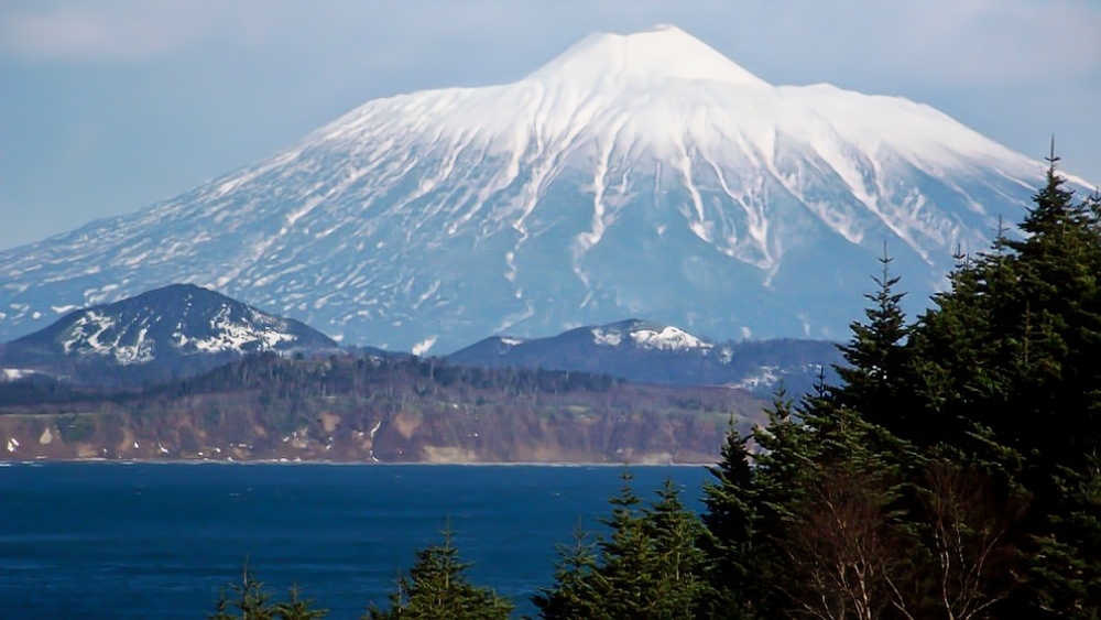 В кадре — действующий вулкан Тятя (1819 м) с белоснежной вершиной, окруженный изумрудными лесами. На побережье — скалистые утесы с арками и гротами, выточенные океаном. Вода Охотского моря переливается оттенками синего и бирюзового. Координаты: 44°00′ с.ш. 145°30′ в.д. Температура воздуха +12°C, воды +6°C. Видны стаи морских птиц и следы китов у берега.