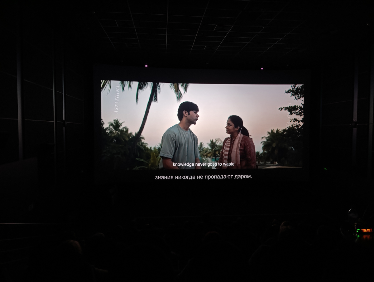 Цветная фотография горизонтальной ориентации. Тёмный кинозал. На экране кадр из фильма «Шрикант». Парк, пальмы, бананы, голубое небо. В центре кадра слева, полубоком, Шрикант – молодой человек, тёмные волосы, стрижка, изображён по пояс, в серой футболке. Выражение лица слегка задумчивое. Справа от Шриканта молодая женщина, индианка, показана по пояс. Тёмные волосы собраны в хвостик позади головы, в ушах небольшие серёжки-гвоздики. Она в бордовой блузке с белыми вертикальными полосками и похожем по оттенку тонком шарфе с поперечными полосками. Внимательно смотрит на Шриканта, чуть улыбается. На тёмном фоне внизу белым шрифтом даны субтитры «знания никогда не пропадают даром», выше надпись на английском «knowledge never goes to waste».