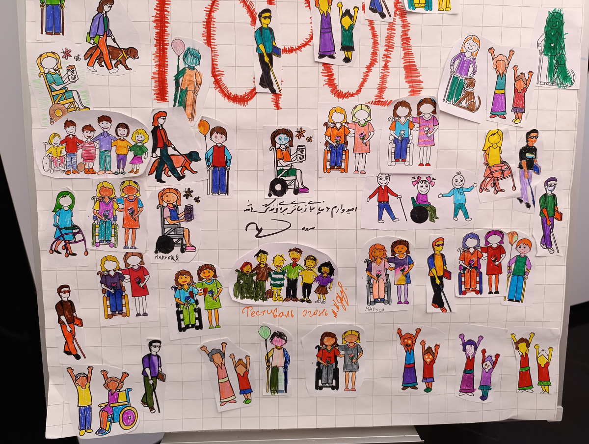 Цветная фотография горизонтальной ориентации. Нижняя часть плаката «Инклюзивный город». На белом листе с крупными клетками наклеены вырезанные из бумаги фигурки людей с инвалидностью и без. Парни с белой тростью, девочки на инвалидных колясках, женщины с ходунками, женщина с собакой-проводником, дети и взрослые с поднятыми вверх руками. Они раскрашены разноцветными фломастерами, какие-то аккуратно, какие-то небрежно. Внизу надпись оранжевым фломастером «Фестиваль огонь» и чья-то подпись. Чуть выше надпись нерусскими буквами.