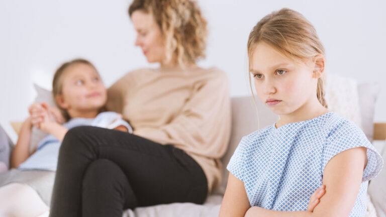    Young upset girl with her arms crossed and blurry mom favoring her twin sister in the background