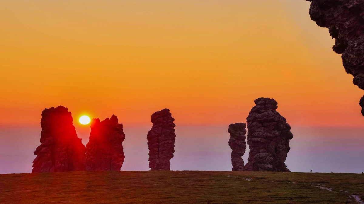 Семь каменных исполинов высотой до 42 метров возвышаются над тундрой, их силуэты окрашены в розово-золотые тона первыми лучами солнца. На переднем плане — моховая тундра с клюквой и брусникой. Координаты: 65°07′ с.ш. 59°58′ в.д., Республика Коми. Высота плато — 420 м над уровнем моря. Температура воздуха -2°C, влажность 80%