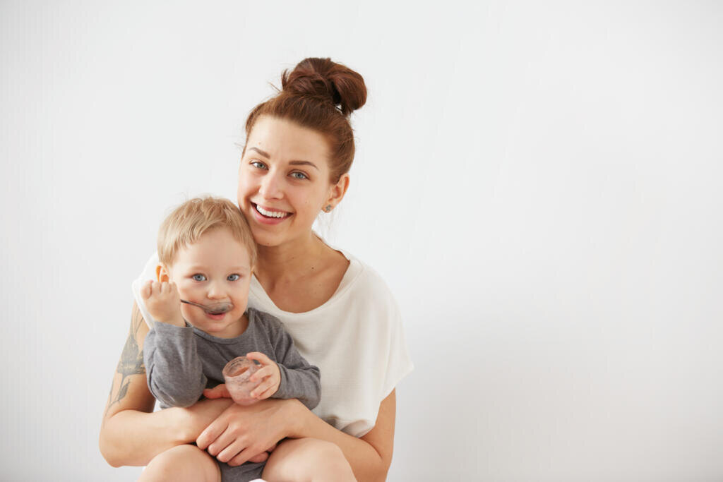    Young attractive mother with her one years old little son dressed in pajamas. Boy eating a fruit smoothie himself in the bedroom at the weekend together, warm and cozy scene. Selective focus. Журналист