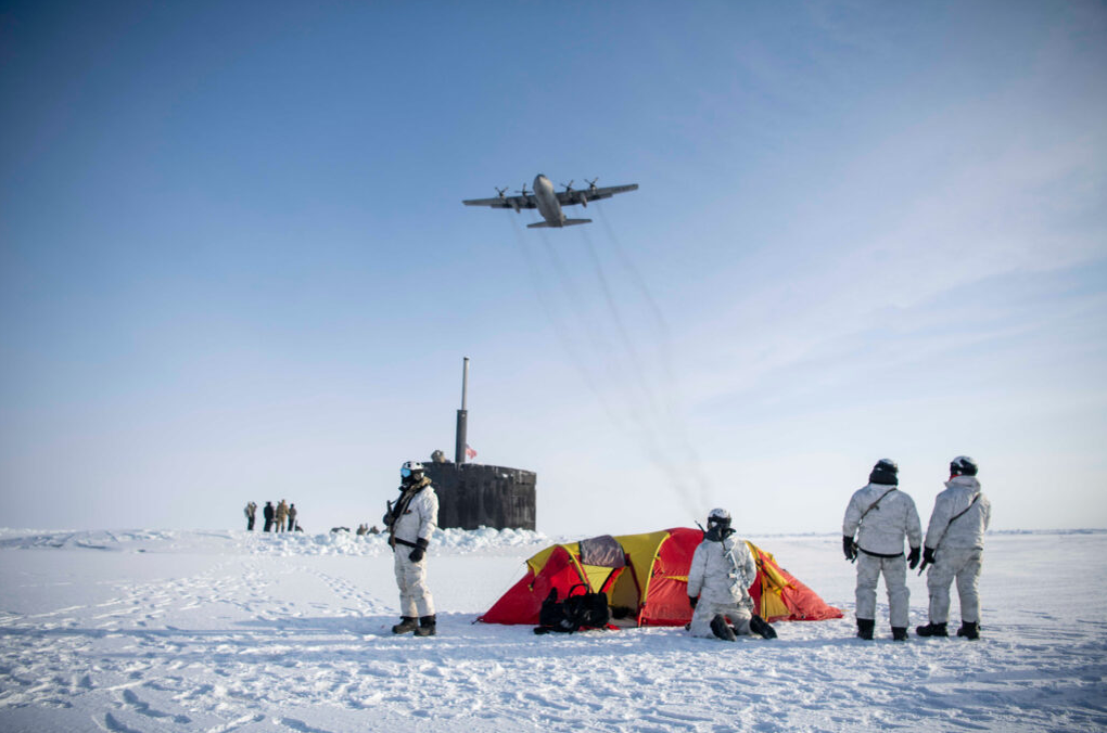 Фото: Самолет ВВС США С-130 Hercules пролетает над группой морских пехотинцев ВМС США, спецназовцев ВМС Норвегии и ударной подводной лодкой USS Hampton во время учений Arctic Edge в Северном Ледовитом океане 9 марта 2024 года. (Источник – Министерство войны США)