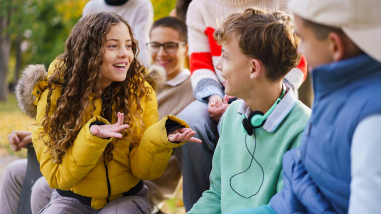    A happy group of teenagers are sitting on a bench and talking in a park during an autumn day