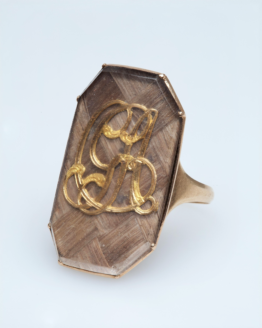 Золотое с гладким ободком. В большом восьмиугольном щитке под стеклом на волосяном фоне, сплетенном шашками, золотая монограмма из букв «CGA». Россия. Конец XVIII в. Золото, стекло, волосы. Размер: 3,1×2,1×2,1 см