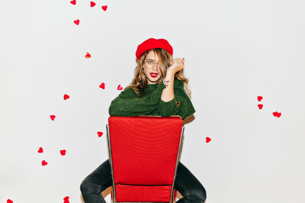    Amazing girl with confident face expression sitting on red chair. Wonderful french woman in green sweater posing on white background in valentine's day. Журналист