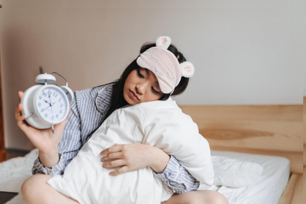    Girl in pink sleeping mask is hugging pillow and looking at alarm clock with sadness Журналист