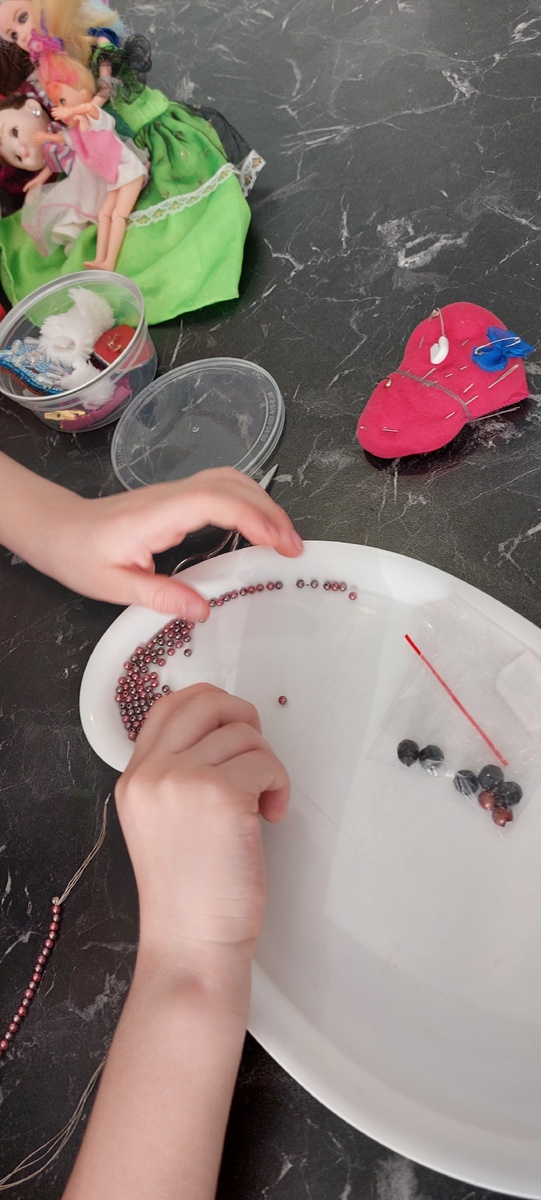 Внучка настойчиво собирает бусины на ожерелье кукле. Собирает на иглу и на сложенную вчетверо особо прочную нить ТИТАН для бисерной вышивки.