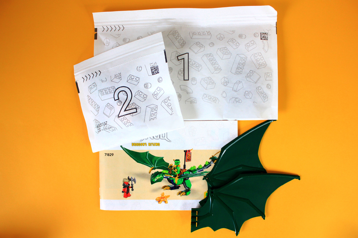 Высыпаем содержимое, и нас встречают два бумажных пакета, два огромных зелёных крыла и инструкция. Это мой первый набор с бумажными пакетиками, и скажу честно, что пластиковые пакеты мне нравятся больше, потому что через них я любил рассматривать ещё не распакованные детальки. Инструкция как и обычно в последние несколько лет не повторяет дизайн с коробки, а представляет набор на нейтральном фоне. Кто знает, может бумажные инструкции тоже скоро уйдут в прошлое, как и пластиковые пакеты ради экологичности или экономии.