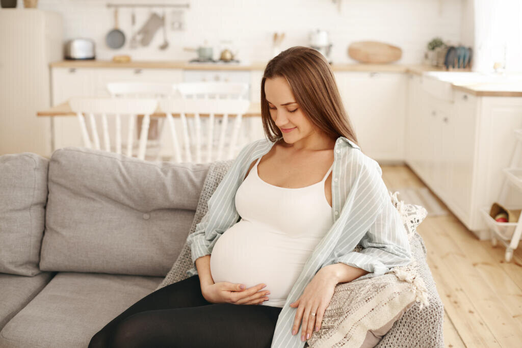    Horizontal shot of beautiful young Caucasian expectant mother communicating with her unborn child, talking to him and stroking her tummy, feeling happy and relaxed, sitting on sofa at home Журналист