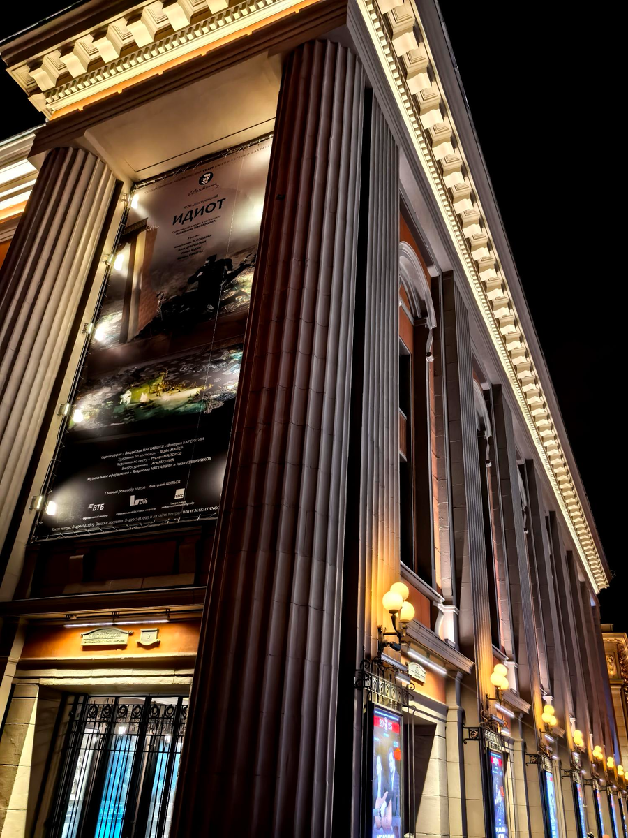 Вечерний город зажигает огни, и величественное здание театра предстаёт во всей своей красоте. Высокие колонны, мягкое золотое освещение и афиши у входа создают атмосферу предвкушения — здесь начинается мир искусства, эмоций и глубоких историй.