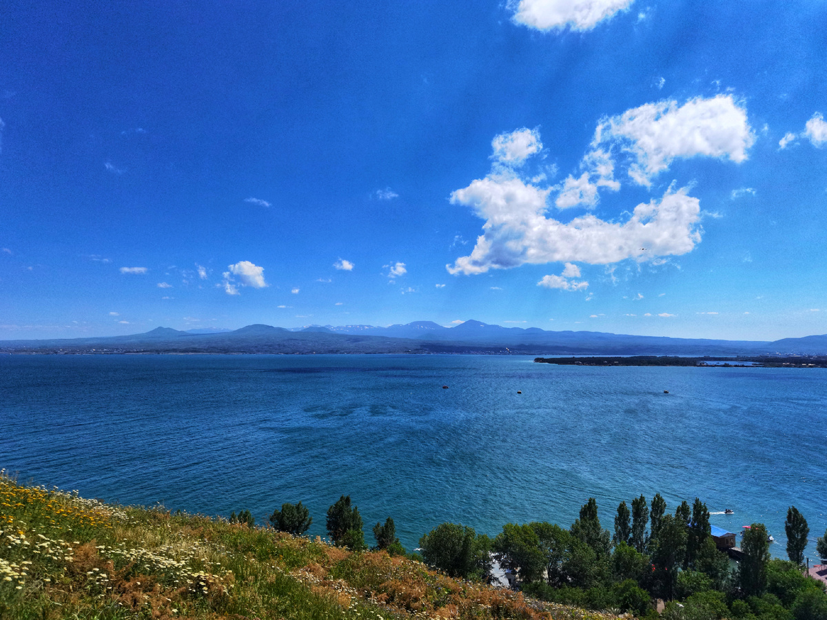 Широкая гладь озера уходит к горизонту, где синие горы поднимаются мягкой линией, словно древняя стража этих вод. Ветер тихо касается поверхности, небо рассыпает над ней лёгкие облака — и всё вокруг дышит спокойствием. Здесь время будто замедляется: остаётся только простор, свет и редкое чувство настоящей свободы.