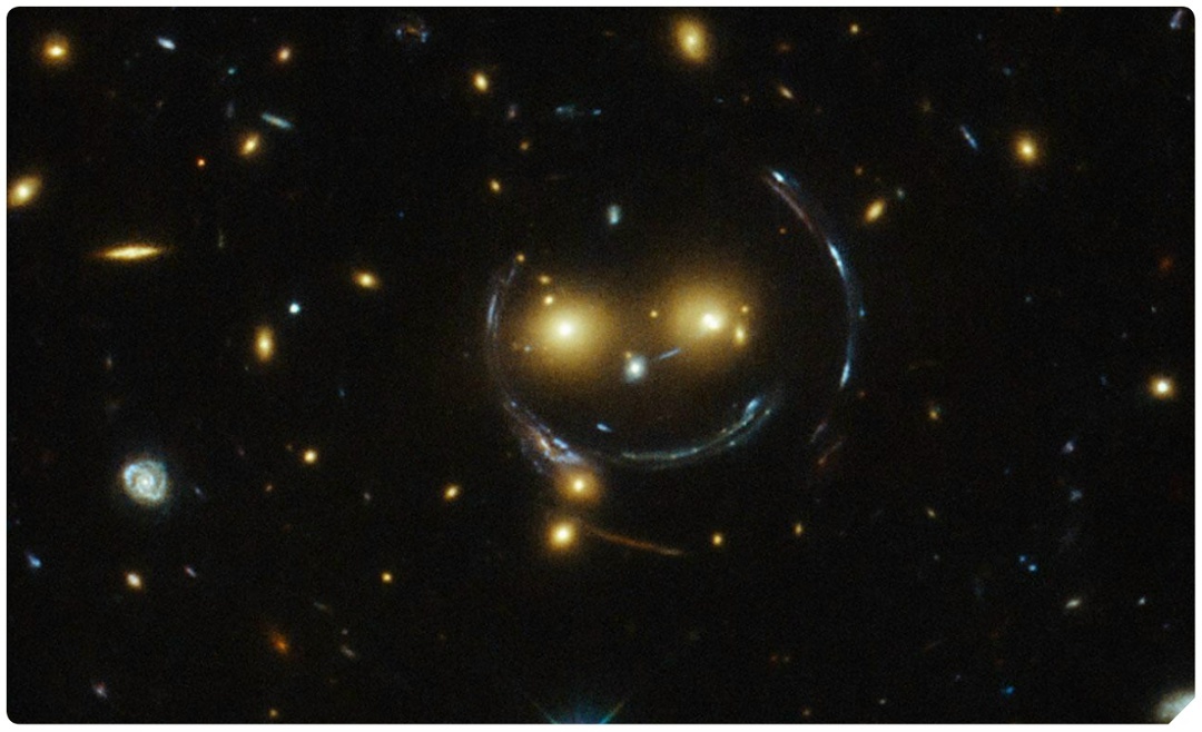 Рис.2. Искажение формы галактик из-за гравитационной линзы делает снимок похожим на смайлик—так его и называют. Свет от далёких галактик искривляется при прохождении мимо скопления галактик—двух ярких галактик—«глаза смайлика»