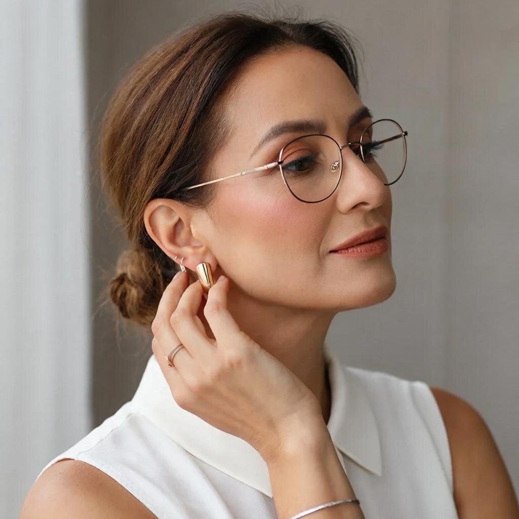 Женщина 40+ в белой блузке показывает гладкие серьги-капли у мочки, аккуратный блеск делает лицо собраннее