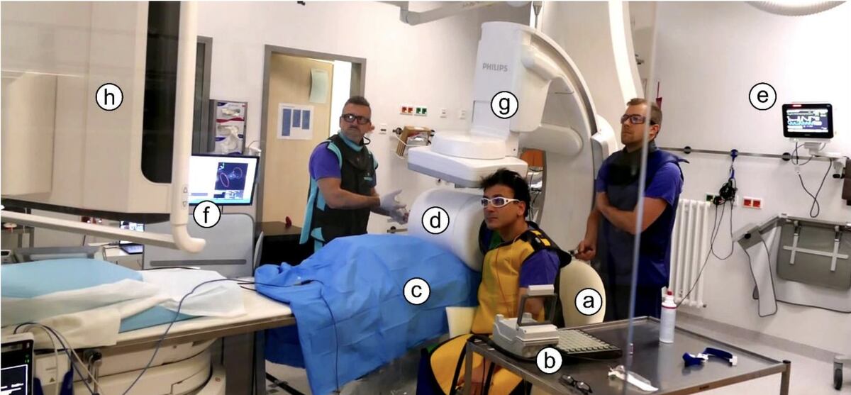    Ангиографическая лаборатория, где проводился эксперимент. a — кресло добровольца; b — устройство ввода/инъекции; c — рука добровольца в сканере; d — MPI-сканер; e — монитор жизненных показателей; f — оборудование и интерфейс управления сканером; g — рентгеновская система для ангиографии; h — экран с изображением ангиографии ./ © Patrick Vogel (2026)