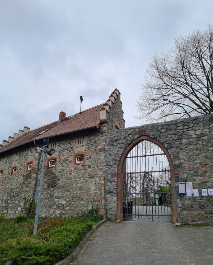 в центре снимка находится высокая стрельчатая арка ворот, закрытая металлической решеткой. Слева к воротам примыкает двухэтажное каменное здание с характерной зубчатой крышей из красной черепицы. Фасад здания сложен из необработанного камня, а небольшие окна обрамлены красноватым песчаником