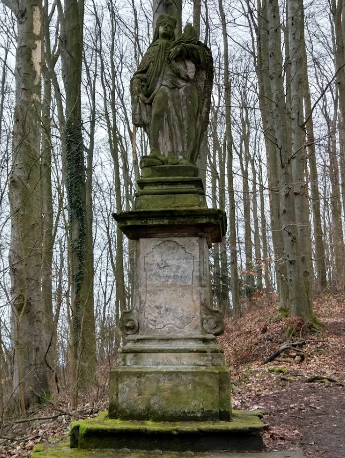 На фотографии запечатлена барочная скульптура святой Людмилы Чешской, расположенная на лесной тропе по пути к замку Гоуска. Этот памятник является не только объектом искусства, но и важным символом для верующих и любителей мистики.