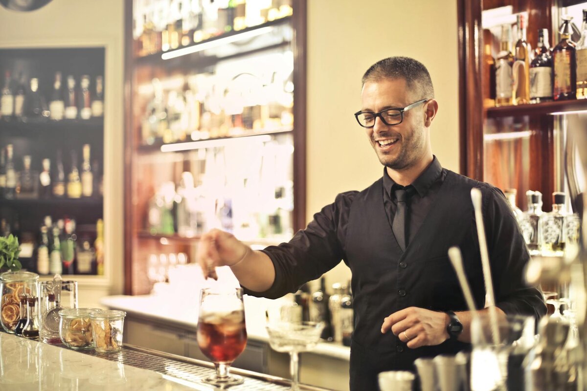 Happy young male barkeeper standing at counter and preparing alcohol cocktail for order while workin