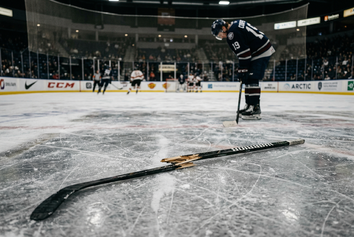 Сломанная хоккейная клюшка на льду, видно место перелома шафта, на фоне размыто стоит хоккеист с опущенной головой, ледовая арена