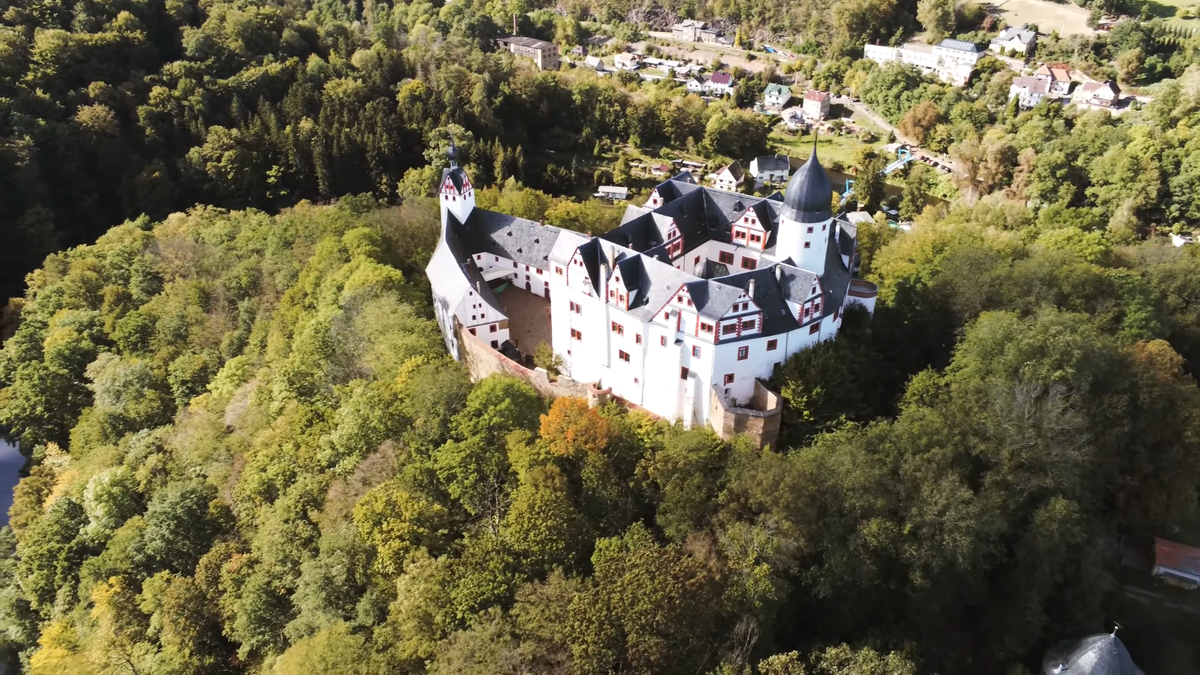 Сказочный замок на скале в Германии. Белые стены старинной крепости контрастируют с золотой и зеленой листвой деревьев на фоне панорамы саксонских полей.