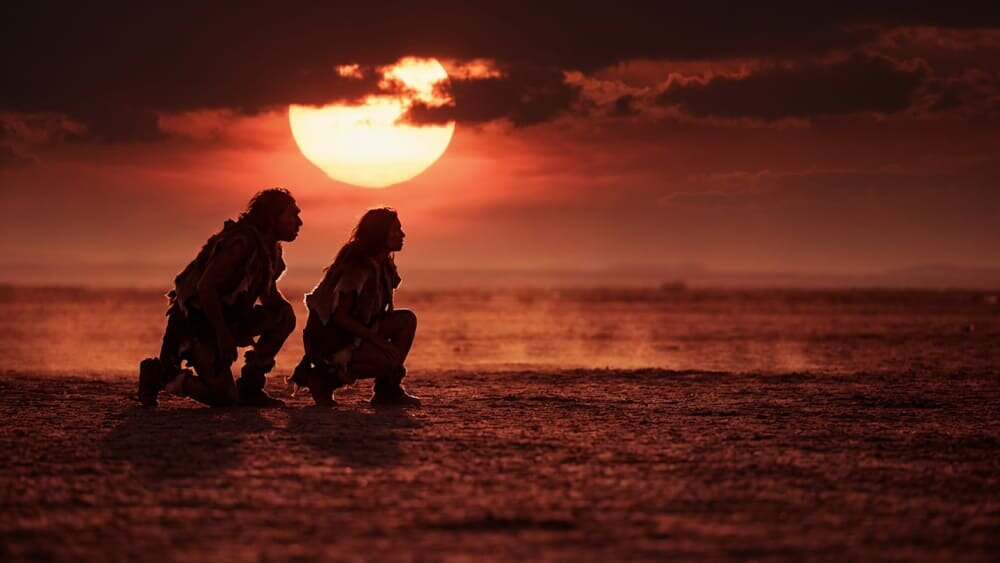 На фотографии изображены два древних человека в шкурах, сидящих на корточках на равнине перед огромным заходящим солнцем и красным небом с темными облаками на горизонте. Изображение сгенерировано в ИИ для иллюстрации идеи.