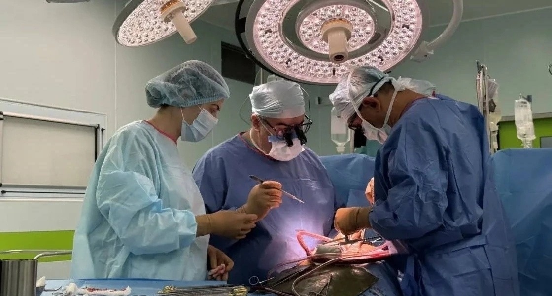    Три хирурга в голубых халатах работают в операционной под ярким светом над инструментами Главный редактор