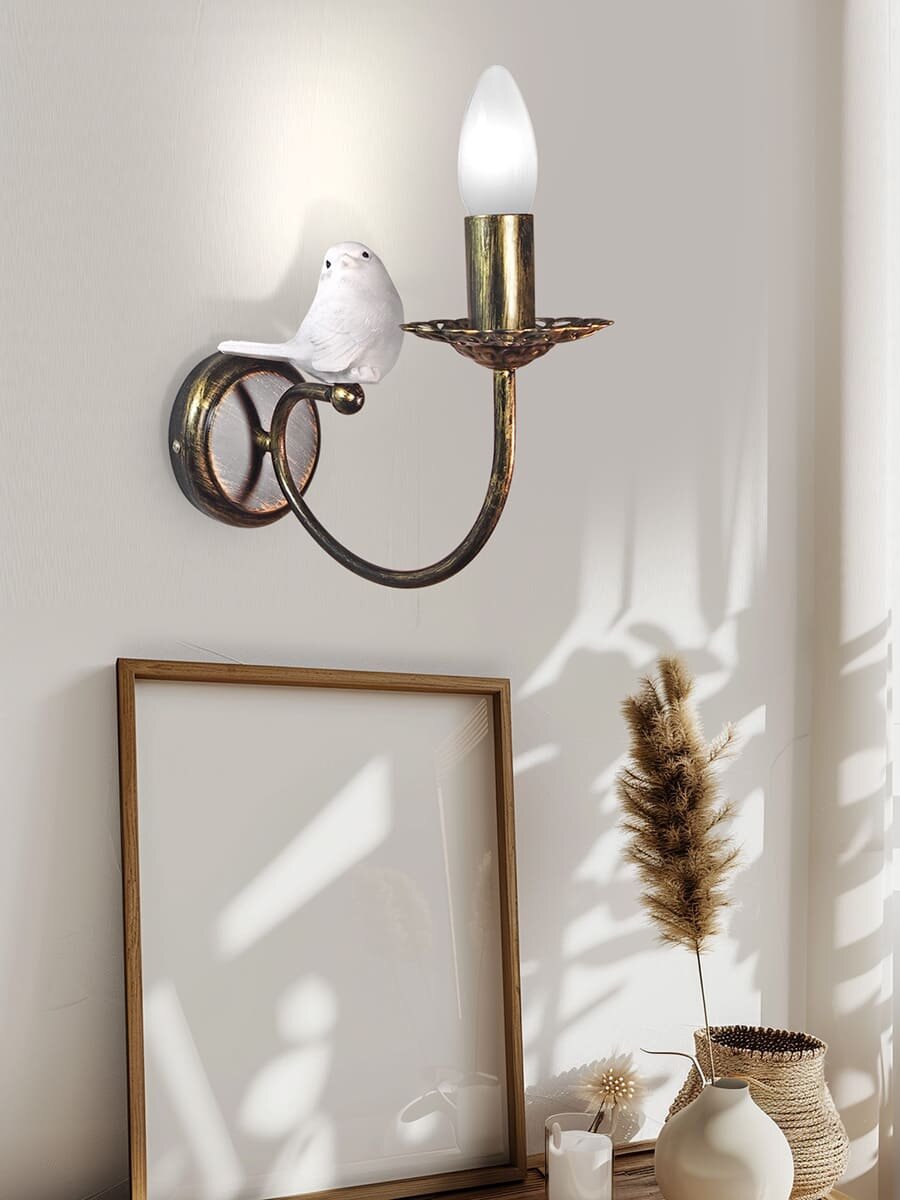 Бра с птичками в S1298-1 Петрасвет: Элегантный полет фантазии в вашем интерьере
Освещение в интерьере — это не просто функциональная необходимость, но и мощный инструмент создания атмосферы. Одним из самых изящных способов добавить помещению уюта и индивидуальности являются настенные светильники. Сегодня мы хотим познакомить вас с настоящей жемчужиной ассортимента нашего каталога — Бра с птичками S1298-1 Петрасвет. Этот светильник — идеальный пример того, как осветительный прибор может стать ключевым декоративным акцентом, сочетая в себе художественную выразительность и высокое качество российского производства.

Дизайн, вдохновленный природой
Модель бра с птичками на стене S1298-1 привлекает внимание в первую очередь своим оригинальным художественным решением. Это не просто плафон на подставке, это миниатюрная скульптурная композиция. Две стилизованные птички, замершие в грациозном движении, словно только что присели отдохнуть на ветку или край гнезда. Их силуэты проработаны с большой любовью к деталям, что превращает обычный светильник в объект искусства.

Ключевой элемент дизайна — это цветовое решение. Основной цвет корпуса — благородный черный, который всегда выглядит стильно и графично. Он отлично контрастирует со светлыми стенами и добавляет глубины темным поверхностям. Но настоящая "изюминка" — это покрытие цвет черный с патиной золота. Искусственно состаренное покрытие с проступающими золотистыми нотками создает эффект антиквариата, придавая изделию особый шарм и визуальную теплоту. Золотая патина выгодно подчеркивает рельеф фигурок птиц, делая их более выразительными и объемными. Этот светильник прекрасно впишется в интерьеры в стиле классика, ар-деко, прованс или современную неоклассику.

Идеальные пропорции: компактность и выразительность
При создании настенных светильников важно найти баланс между декоративностью и функциональностью. 