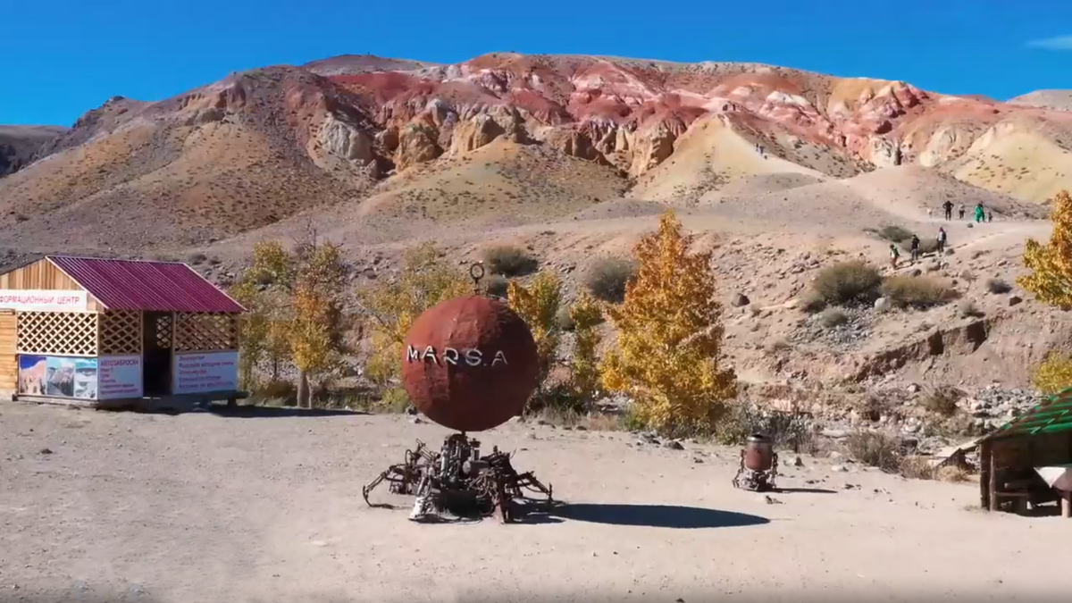 «Вход на территорию легендарных Марсовых гор (Кызыл-Чин). У подножия цветных холмов туристов встречает необычный арт-объект в виде планеты Марс на металлическом основании. Здесь начинается путешествие в доисторические времена: на склонах гор, окрашенных железом в красные и охристые тона, до сих пор можно найти следы древнего океана Тетис. Осень на Алтае добавляет золота лиственниц в и без того невероятную палитру»