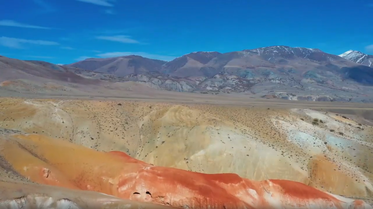 «Где-то на границе миров: разноцветные склоны долины Кызыл-Чин, более известные как "Алтайский Марс". Невероятная палитра от огненно-рыжего до нежно-бежевого создана самой природой миллионы лет назад. На заднем плане — величественные заснеженные вершины Курайского хребта под пронзительно синим небом Кош-Агачского района».