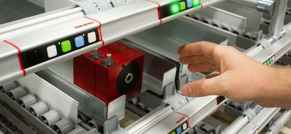 Pick-by-Light (световая комплектация).На изображении показан стеллаж с установленными на ячейках свето-цифровыми модулями. Каждый модуль включает LED-кнопку, которая подсвечивается определённым цветом (например, зелёным, красным или другим, в зависимости от настроек системы), и цифровое табло, отображающее количество товара, которое необходимо отобрат