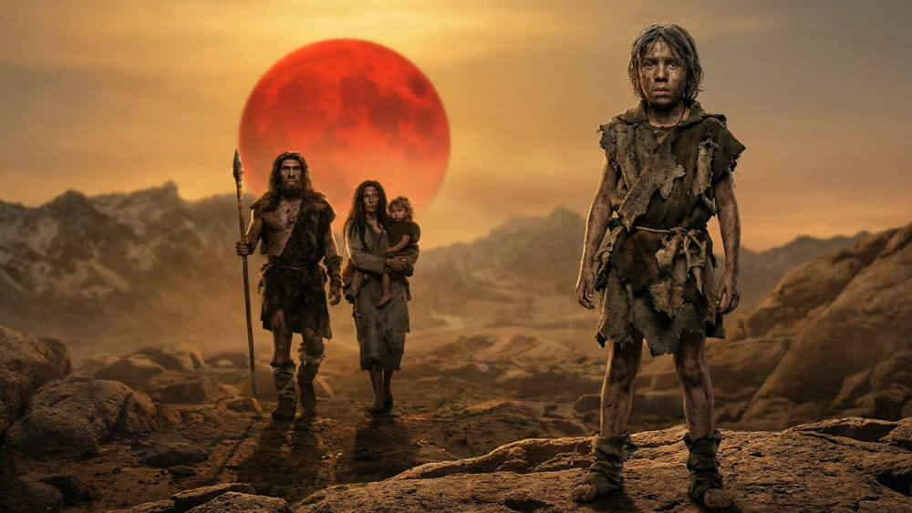 На фотографии изображены древние люди в изношенных шкурах, идущие по каменистой пустынной местности на фоне огромного красного солнца и гор под оранжевым небом. Изображение сгенерировано в ИИ для иллюстрации идеи.