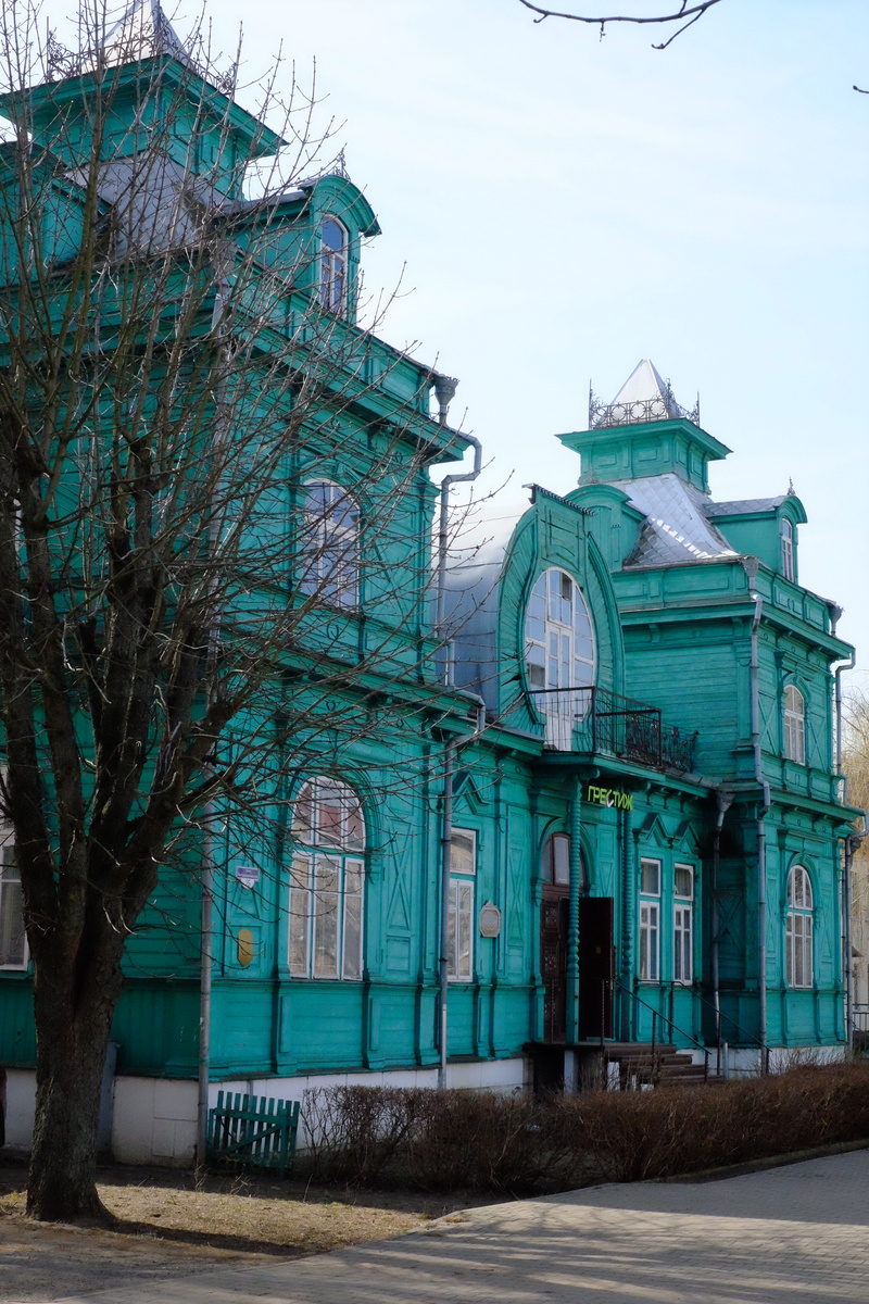 Деревянное здание в форме буквы «Г» с двускатной крышей. Главный фасад — с двумя башнями-шатрами. А в центре — круглая мансарда и балкон. Фото автора.