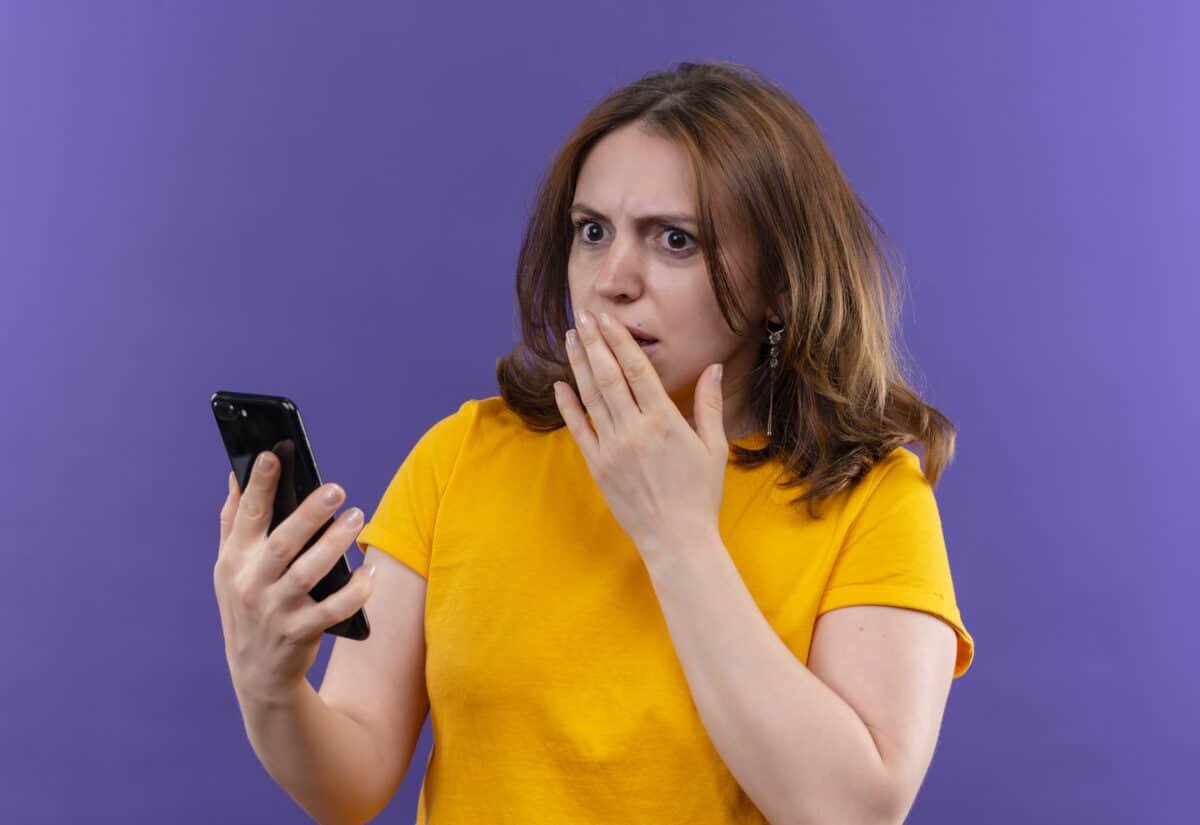    surprised young casual woman holding mobile phone with hand on mouth on isolated purple background Александр Петров