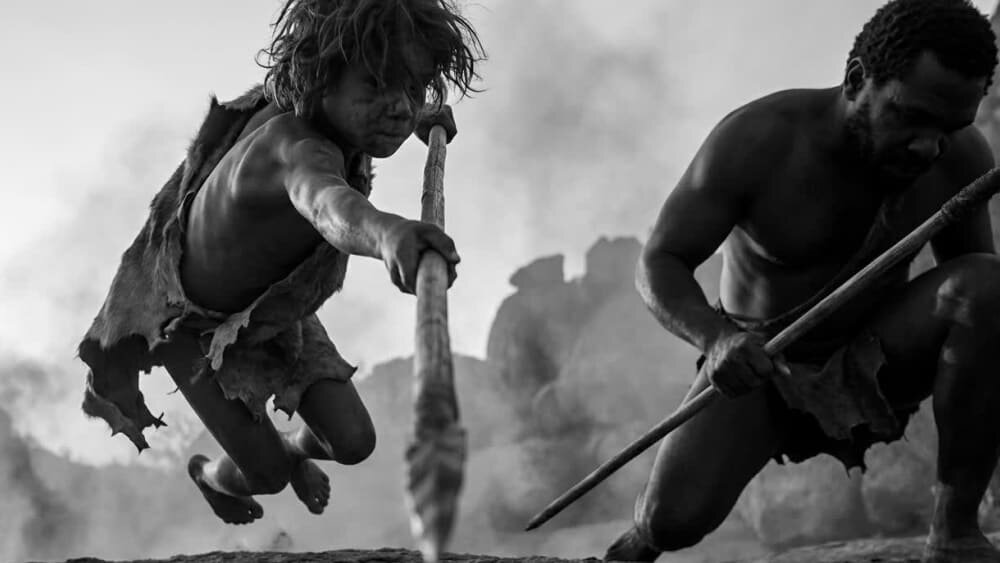 На фотографии изображены ребенок и мужчина с копьями, атакующие в прыжке на фоне скал и дыма. Одеты в шкуры, черно-белый кадр передает динамику. Изображение сгенерировано в ИИ для иллюстрации идеи.