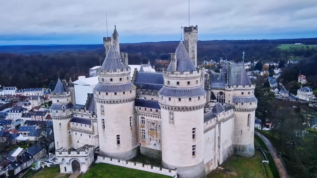 Панорамный вид на замок Пьерфон (Château de Pierrefonds) во Франции с высоты птичьего полета. Величественные башни, мощные стены и окружающий город Пьерфон под пасмурным небом.
