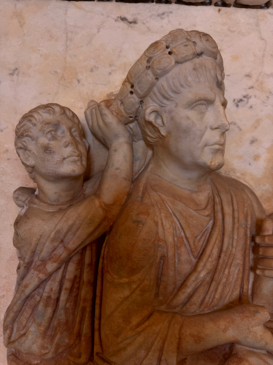 Император с венком на голове и рядом маленькая фигура сзади, которая тянется к уху. Это и есть тот самый момент — шёпот в ухо