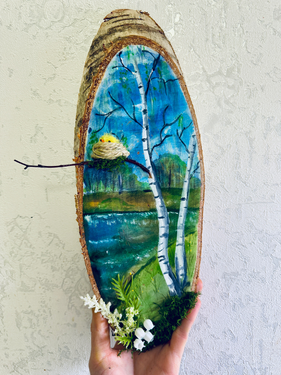 Сегодня такие деревянные спилы с росписью очень популярны. Они красиво смотрятся в интерьере, подходят для подарка и становятся интересной творческой работой для детей и взрослых.
Для основы берут деревянный спил овальной или круглой формы. Поверхность нужно хорошо зачистить наждачной бумагой, чтобы она стала гладкой и удобной для рисования. После этого спил желательно покрыть светлой грунтовкой или белой акриловой краской. Так рисунок будет ярче и аккуратнее.
Когда основа высохнет, можно приступать к фону. Сначала наносят небо, воду, траву или лес вдали. Затем постепенно прорисовывают главные детали: деревья, берег, листья, цветы. Для такой работы удобно использовать акриловые краски, потому что они быстро сохнут и хорошо ложатся на дерево.
Чтобы работа выглядела объёмнее и интереснее, можно добавить декор. Например, приклеить маленькую веточку, мох, гнездо из шпагата, искусственные цветы или другие природные элементы. Именно такие детали делают спил особенно уютным и необычным.
После полного высыхания готовую работу лучше покрыть лаком. Это защитит краску и поможет сохранить изделие надолго.
Такой расписной спил — это не просто поделка, а маленькая картина на дереве, которая создаёт весеннее настроение и радует глаз.
