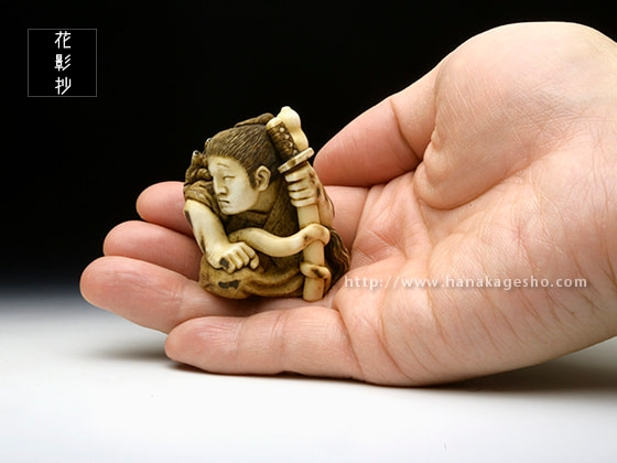 Нэцкэ «Записи о призрачных явлениях в доме Ино:». Автор: КОМА (狛). Материал: рог оленя и чёрное дерево. Третий год эпохи Рэйва (2021). Вид спереди и демонстрация размера.