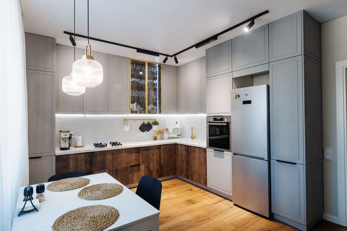 На фото представлен интерьер кухни, который идеально иллюстрирует тренд на «современную классику» (transitional style). Это пространство, где строгие линии встречаются с уютом, а практичность не конфликтует с эстетикой. Дизайнеру удалось создать комнату, которая выглядит дорого и статусно, но при этом остается очень жилой и гостеприимной.
Давайте разберем ключевые приемы, которые делают этот интерьер таким гармоничным, и узнаем, как применить их в своей квартире.
1. Двухцветный гарнитур: Игра на контрастах
Главная фишка этой кухни — разделение цветов по горизонтали. Это смелое, но беспроигрышное решение.
Верх — светлый: Шкафы верхнего ряда выполнены в оттенке «пыльный серый» (или светло-бежевый тауп). Матовая поверхность и классическая филенка (стиль Shaker) добавляют воздуха и легкости.
Низ — темный: Нижние фасады отделаны под темное дерево (орех или венге). Это «заземляет» интерьер, делает его устойчивым и визуально более дорогим. К тому же, темный низ практичнее — на нем меньше видны случайные брызги или следы касаний.
Белая столешница и фартук: Они работают как разделитель и светоотражатель, не давая темному низу «съесть» пространство.
2. Освещение: Сценарии для разного настроения
Свет в этой кухне продуман на нескольких уровнях, что превращает рутинную готовку в приятный процесс.
Трековая система на потолке: Черный трек с поворотными спотами — это современное решение. Он дает мощный направленный свет, который можно перенаправить куда нужно: на рабочую зону, на мойку или на холодильник.
Акцентные подвесы над столом: Два крупных светильника с ребристым стеклом и латунным основанием создают уютную зону для ужина. Они визуально «опускают» потолок над столом, делая пространство более камерным.
Подсветка рабочей зоны: Под верхними шкафами пущена LED-лента. Это критически важно для удобства: вы не стоите в собственной тени, когда режете продукты.
📍 Приходите знакомиться:
г. Омск, ул. Фрунзе 1/4, ТЦ «Миллениум», 11 этаж
📞 Звоните или пишите: +7 (3812) 99-20-38