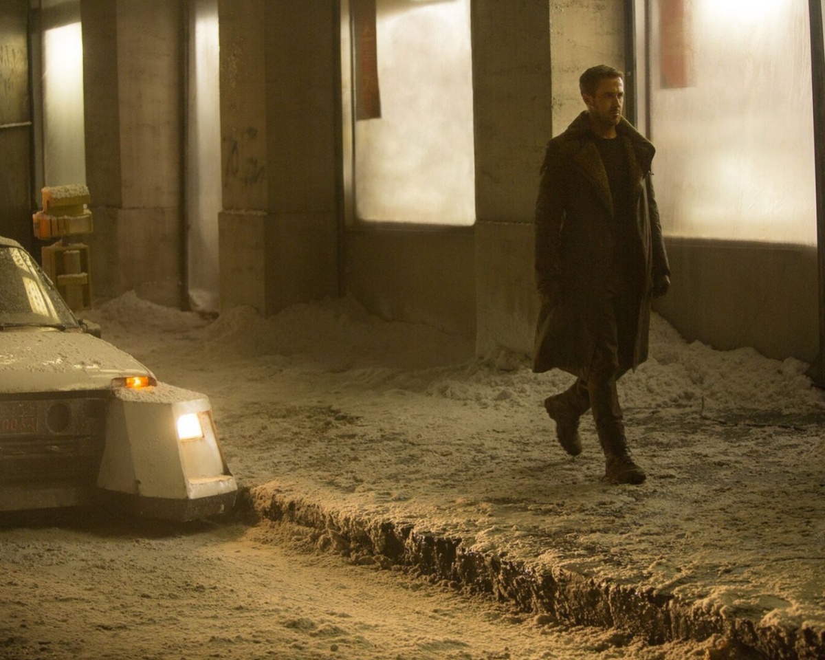 Кадр из фильма «Бегущий по лезвию 2049» (реж. Дени Вильнёв, 2017) Офицер Кей (Райан Гослинг) идет на фоне гигантской неоновой фонарей. Его силуэт чётко выделяется на фоне унылого города. Почему этот кадр важен? Он показывает, как свет в фильме не прячет грязь, а подчёркивает её: каждый неоновый блик проявляет влажную фактуру асфальта, каждое отражение делает видимой тонкую плёнку городской гари на поверхности луж.