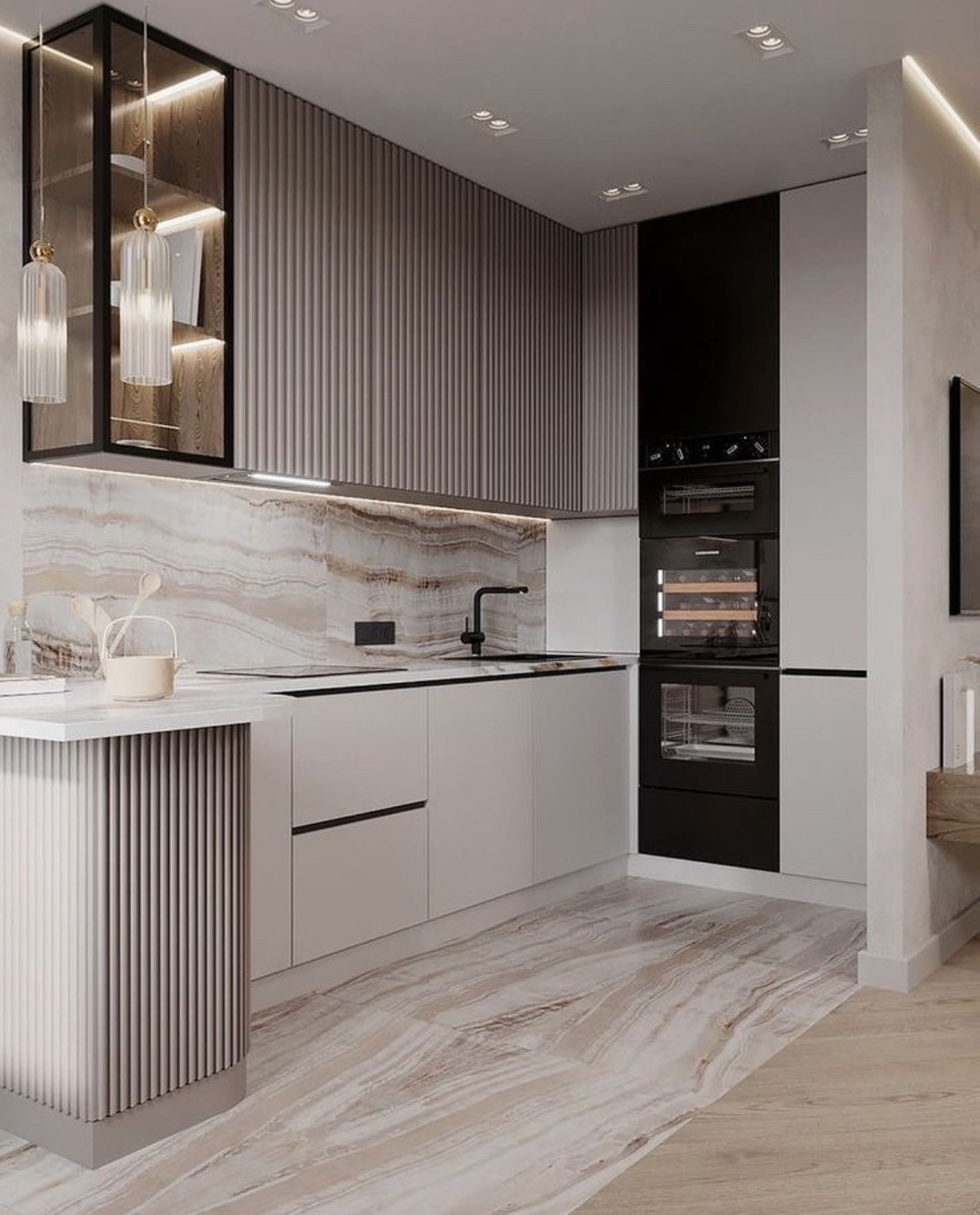На фотографии представлен интерьер, который можно назвать эталоном современного «мягкого минимализма» с нотками премиальной эстетики. Это пространство не кричит о своей дороговизне через вычурный декор или кричащие цвета. Напротив, оно говорит о статусе через безупречное качество материалов, сложную работу с фактурами и выверенный баланс света и тени.
Давайте разберем ключевые дизайнерские приемы этого проекта, которые превращают обычную кухню в произведение искусства, и узнаем, как использовать их в своем доме.
1. Игра фактур: Вертикальный ритм против гладкости
Главная особенность этого интерьера — мастерское сочетание разных поверхностей. Дизайнер избегает монотонности, создавая тактильный конфликт, который радует глаз.
Рейки и фрезеровка: Обратите внимание на верхние шкафы и боковую часть полуострова слева. Они выполнены с вертикальной ребристой фактурой (флейтинг). Этот прием сейчас на пике популярности не просто так: вертикальные линии визуально «поднимают» потолок, делая комнату выше, а рельеф скрадывает мелкие неровности и отпечатки пальцев лучше, чем глянцевый глянец.
Гладкий матовый низ: Нижние фасады — гладкие, матовые, цвета «теплый белый» или «крем». Они работают как спокойный фон, уравновешивая активную фактуру верха.
Камень с характером: Фартук (стеновая панель) и пол выполнены из крупноформатного материала (вероятно, керамогранит под оникс или травертин) с выразительными горизонтальными прожилками. Этот узор добавляет динамики в строгую геометрию шкафов.
2. Цветовая палитра: Сложные нейтральные тона
Здесь нет чистого белого или черного. Дизайнер использовал палитру нюд и тауп.
База: Оттенки песка, кофе с молоком и слоновой кости создают атмосферу уюта и спокойствия. 