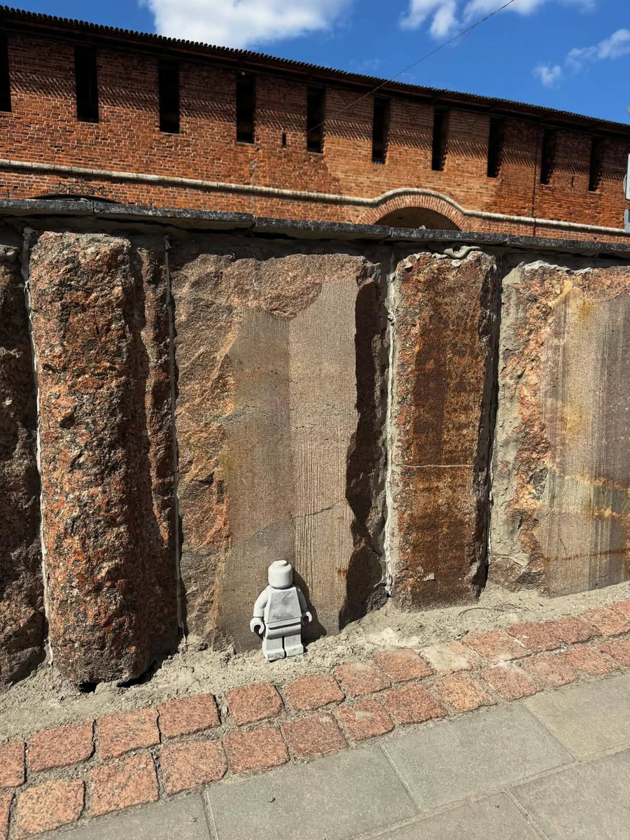 Полный кадр кремлёвской стены: красный кирпич с зубцами поверху, мощные гранитные плиты внизу, выложенная розовым булыжником мостовая — и в самом центре всего этого крошечный бетонный LEGO. Масштаб делает ровно то, чего хотел автор: один маленький человек у большой архитектуры.