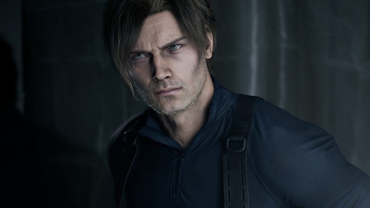    Close-up of a man with blue eyes and stubble, wearing a dark tactical jacket with straps in a dim setting.